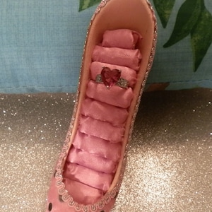 Puede incluir: Porta anillos en forma de zapato rosa con un interior de satén rosa y un anillo con una piedra rosa en el centro.