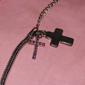 May include: A silver chain necklace with two silver cross charms. One cross is plain and the other is decorated with purple rhinestones.