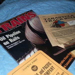 Puede incluir: Una colección de revistas de trenes vintage, incluyendo "Trains" y "Tarantula Track Bulletin". La revista "Trains" presenta una foto de un tren cisterna del Southern Pacific con el texto "Oil Pipeline on Rails". La revista "Tarantula Track Bulletin" presenta una foto de un tren con el texto "Fort Worth & Western".