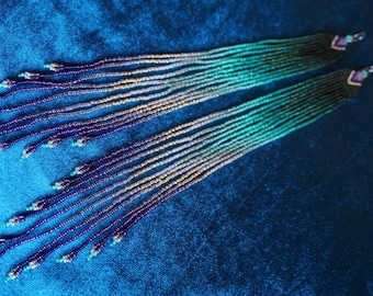 May include: Long beaded earrings with a gradient of colors. The earrings transition from teal to purple, with gold and silver accents. Each earring has a small square top with a purple stone.