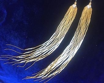 May include: A pair of long, beaded earrings with a gold and white color scheme. The earrings are made of small, round beads that are strung together in a graduated pattern. The earrings are very long and dangle down to the shoulders.