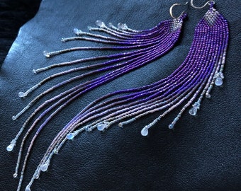 May include: A pair of long, beaded earrings with a gradient of purple and pink colors. The earrings are adorned with small, teardrop-shaped stones.