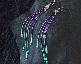 May include: A pair of long, beaded earrings with gold-colored hooks. The earrings feature strands of small, colorful beads in shades of purple, teal, and iridescent hues. The earrings are displayed on a black leather surface.