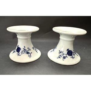 May include: Two white ceramic candle holders with a blue floral design. The candle holders have a wide, flat top and a flared base. The blue design includes a stylized pomegranate and floral elements.