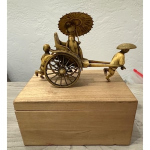 May include: A detailed ivory-colored figurine depicting a rickshaw with a passenger under a parasol, pulled by two figures. The rickshaw sits atop a light-colored wooden box, showcasing intricate craftsmanship and cultural artistry.
