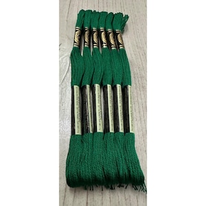 May include: A bundle of emerald green embroidery floss. Each skein is wrapped with a black and white label. The floss is arranged on a light wood surface. The floss is for embroidery, cross-stitch, and other needlecraft projects.