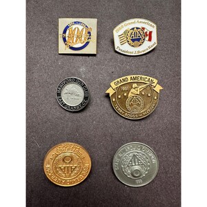 May include: A collection of vintage pins and medallions. The pins feature text and graphics, including "100th Trap & Field", "1968 Grand American", and "Hartford Gun Club". The medallions include "Grand American 1991" and "State All Around Champion 1996".