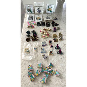 May include: A collection of enamel pins in various shapes and designs, including flags, buildings, and abstract shapes. Some pins are packaged, while others are loose. Colors include green, blue, pink, and gold.