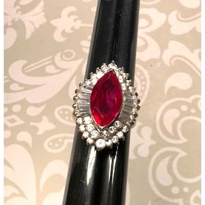 May include: A ring featuring a central marquise-cut red gemstone surrounded by a halo of small round white stones and baguette-cut white stones. The setting is metallic and the ring is displayed on a black tapered stand against a patterned background.