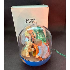 May include: A Christmas-themed snow globe with a clear plastic dome, featuring two children, a dog, and a Christmas tree. The base is blue, and the box behind it has the text "QLX 7279D HOT WHEELS".
