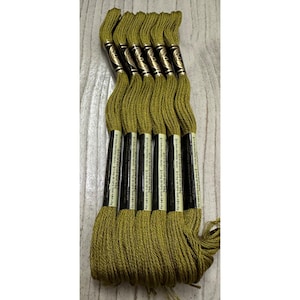 May include: A bundle of olive green embroidery floss. Each skein is wrapped with a black and gold label. The floss is arranged vertically, with the ends of the skeins fanned out at the bottom. The background is a light wood grain.