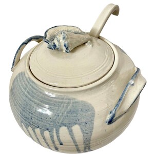 May include: A ceramic soup tureen with a lid and handle, featuring a seashell-shaped knob. The off-white tureen has blue painted accents and a matching ladle. The design is reminiscent of ocean waves.