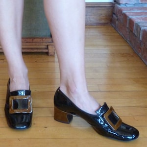 May include: A pair of black patent leather shoes with gold buckles. The shoes have a low block heel.