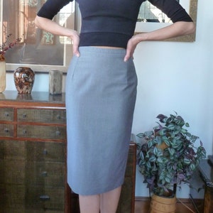 May include: A woman wearing a black mock neck top and a grey pencil skirt. She is standing in a room with a wooden floor and a dresser.