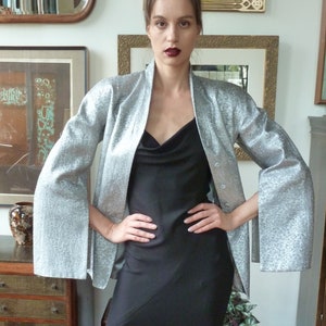 May include: A woman wearing a silver sequined jacket with wide sleeves over a black dress. The jacket has a button closure and the dress has a cowl neck.