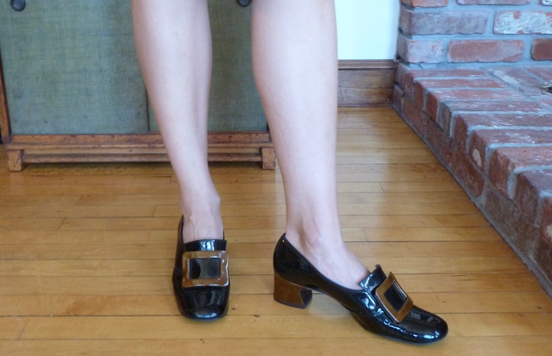 May include: A pair of black patent leather shoes with gold buckles. The shoes have a low block heel.