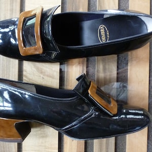 May include: Pair of black patent leather shoes with a brown buckle. The shoes have a low heel and a pointed toe.