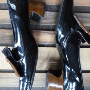 May include: A pair of black patent leather shoes with brown heels. The shoes feature a buckle detail on the front.