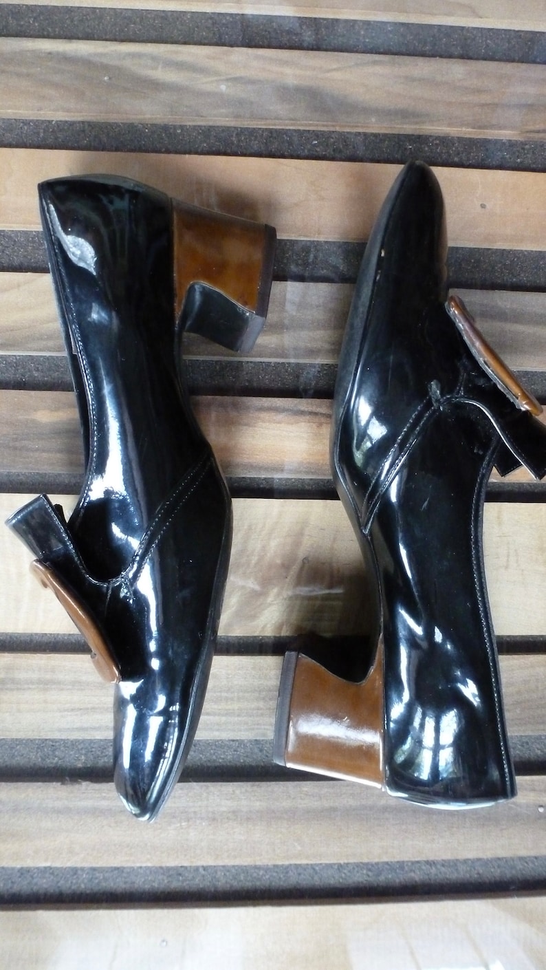 May include: A pair of black patent leather shoes with brown heels. The shoes feature a buckle detail on the front.