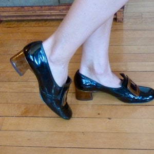 May include: A pair of black patent leather heeled shoes with gold buckles. The shoes have a chunky heel and a pointed toe.