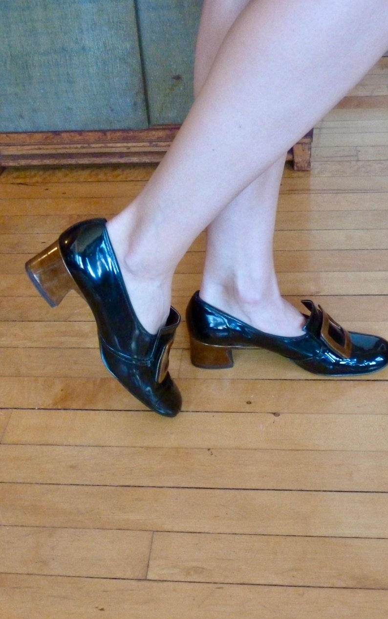 May include: A pair of black patent leather heeled shoes with gold buckles. The shoes have a chunky heel and a pointed toe.