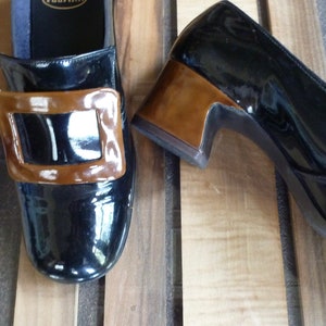 May include: A pair of black patent leather shoes with a brown buckle and a low heel.