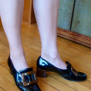 May include: A pair of black patent leather shoes with a brown buckle. The shoes have a chunky heel and a pointed toe.