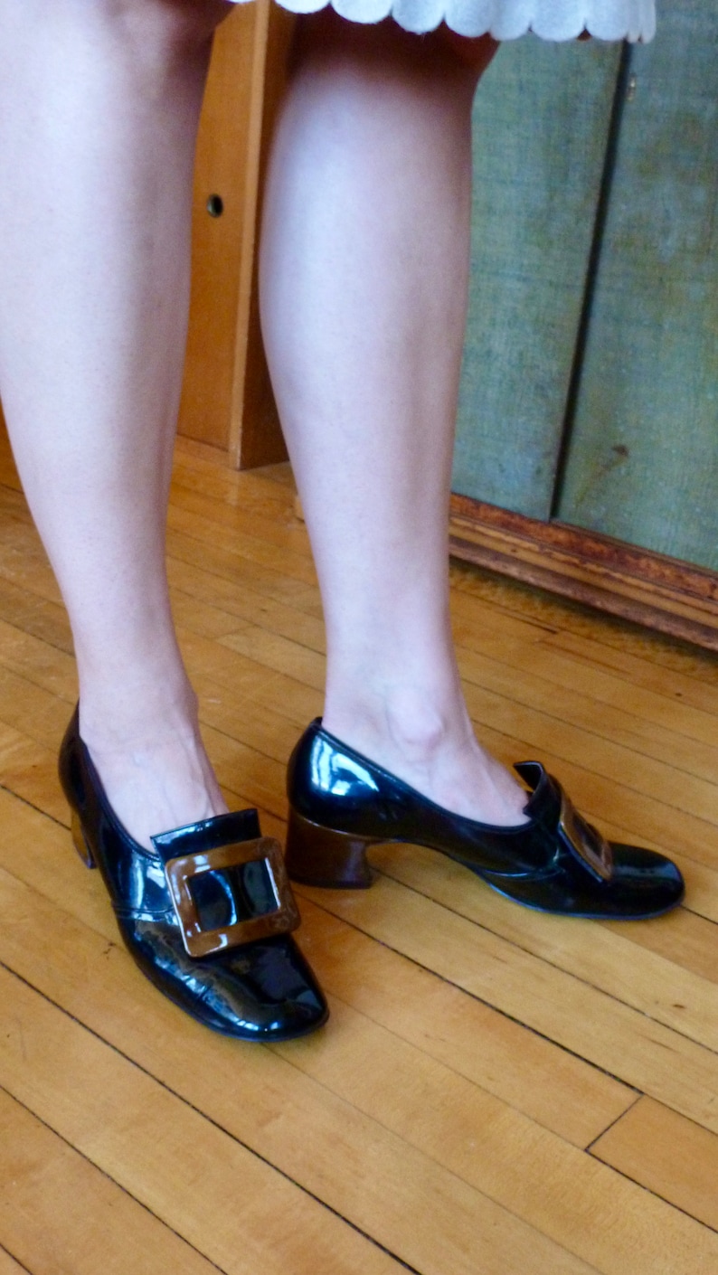 May include: A pair of black patent leather shoes with a brown buckle. The shoes have a chunky heel and a pointed toe.