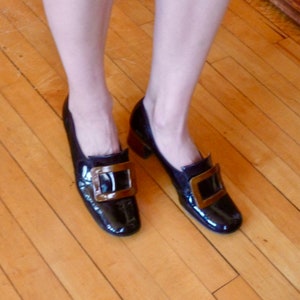 May include: A pair of black patent leather shoes with gold buckles. The shoes have a pointed toe and a low heel.