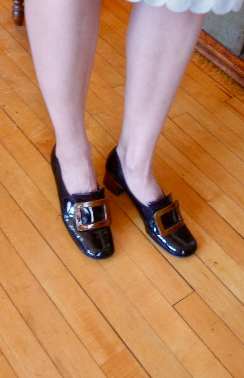 May include: A pair of black patent leather shoes with gold buckles. The shoes have a pointed toe and a low heel.