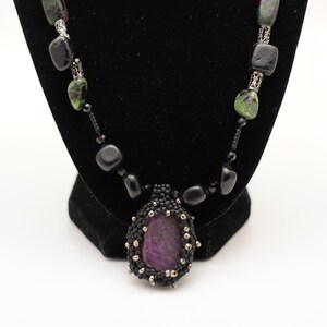 May include: A black beaded necklace with a large purple gemstone pendant. The necklace features black and green stones and silver accents.