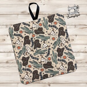 May include: A beige quilted pot holder with a repeating pattern of cacti, cowboy hats, and skulls. The text "Howdy" is repeated throughout the pattern.