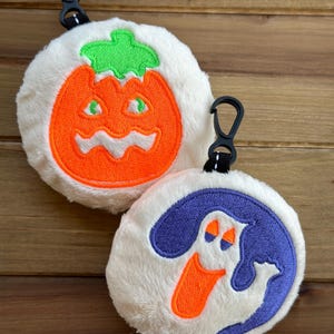 May include: Two white plush Halloween-themed keychains. One features a bright orange pumpkin with a green stem and a spooky face. The other features a purple ghost with a bright orange mouth and eyes.