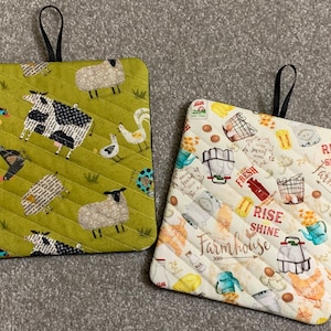 May include: Two square quilted pot holders. One is green with farm animal illustrations, including cows, sheep, and chickens. The other is cream-colored with a farmhouse theme, featuring eggs, milk jugs, and the words "Rise Shine Farmhouse."