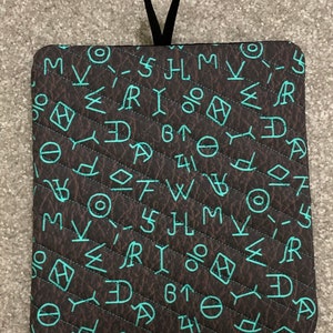 May include: Square, quilted pot holder with a dark brown background and a pattern of turquoise symbols. The pot holder has a black hanging loop at the top. The symbols are in a variety of shapes and sizes.
