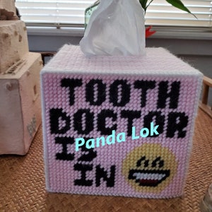 Puede incluir: Tapa de caja de pañuelos de color rosa con bordado negro que dice "Tooth Doctor is in" y una cara sonriente amarilla. La tapa está hecha a mano y tiene un borde blanco.