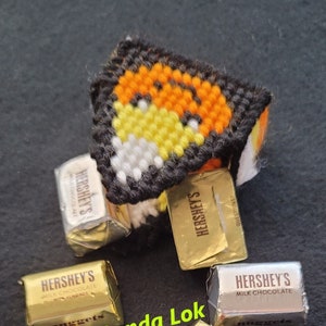 Può includere: Primo piano di cinque barrette di cioccolato Hershey's, con un'artigianato fatto a mano a forma di mais dolce sullo sfondo. Le barrette di cioccolato sono avvolte in foglio d'alluminio dorato e argentato, con il logo Hershey's e le parole "Cioccolato al latte" e "Con mandorle" stampate su di esse. L'artigianato a forma di mais dolce è fatto di filo ed è arancione, giallo e bianco.