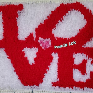 May include: A close-up of a red and white rug with the word "LOVE" spelled out in fluffy yarn. The rug is made on a canvas base and has a grid pattern.