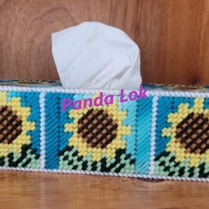 May include: A tissue box cover with a sunflower design. The cover is made of colorful yarn and features three sunflowers with brown centers, yellow petals, and green leaves. The text "Panda Lok" is visible on the cover.