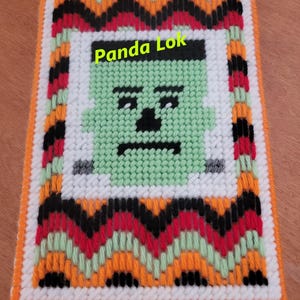 May include: A green and white cross-stitch of a Frankenstein's monster face with a black and white zigzag border. The border is in shades of orange, red, green, and black. The text "Panda Lok" is in the upper center of the image.