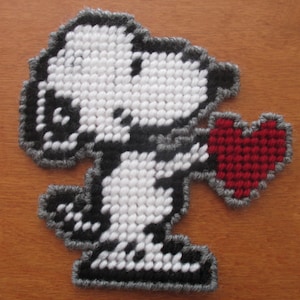 May include: A handmade Snoopy character holding a red heart, crafted from yarn using a plastic canvas technique. The character is primarily white with black accents and a gray border. The heart is a solid red color.