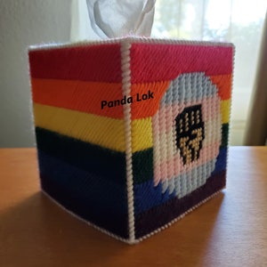 May include: A rainbow-colored tissue box cover with a white border and a pink, blue, and white fist with a gold outline in the center. The text "Panda Lok" is written on the front of the box.