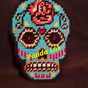 以下が含まれることがあります： 額に赤いバラがついた青い砂糖の頭蓋骨のクロスステッチ。頭蓋骨は黒い目とオレンジ色の歯を持つ白い口をしています。頭蓋骨には「Panda Lok」という文字が見えます。