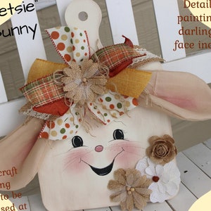 May include: A white wooden cutting board with a painted bunny face, brown fabric ears, and a burlap bow with orange and green polka dot ribbon. The text "Betsie" Bunny and "PDF" Pattern Details for painting this darling little face included!" is visible. The text "Small craft cutting board to be purchased at Walmart!" and "Old Road Primitives" is also visible.