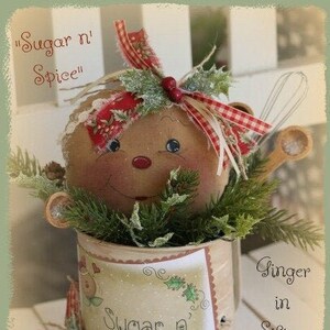 May include: A handmade Christmas decoration featuring a gingerbread person doll with a red and white gingham bow, nestled in a vintage-style tin can with a label that reads "Sugar n' Spice". The tin can is decorated with holly leaves and berries, and the doll is surrounded by greenery.