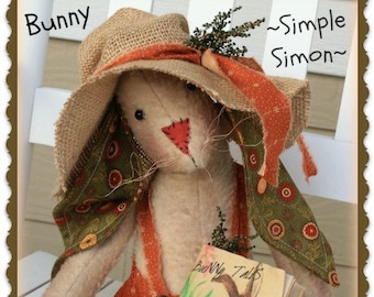 May include: A handmade bunny doll wearing a straw hat with an orange ribbon and a green and orange floral print dress. The doll is holding a book titled "Bunny Tails".