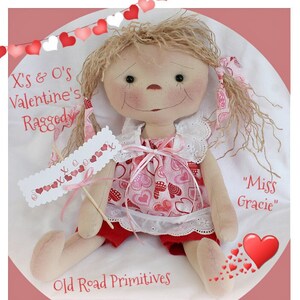 Könnte beinhalten: Eine handgemachte Stoffpuppe mit blonden Haaren, die ein rotes und pinkfarbenes Kleid mit Herzmuster und einen weißen Spitzenkragen trägt. Die Puppe hält ein Schild mit der Aufschrift "X's & O's Valentine's Ragged" und ist mit "Miss Gracie" beschriftet.