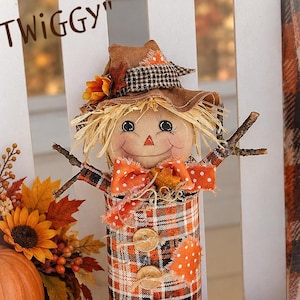 May include: A decorative scarecrow figure named Twiggy, crafted from a tin can covered in orange, black, and white plaid fabric. It features a fabric hat, twig arms, a painted face, a polka-dot bow, and three wooden buttons on the front.