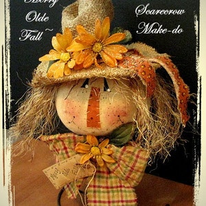 May include: A handmade scarecrow figurine with a burlap hat, orange and yellow flowers, and a plaid fabric base. The scarecrow has a painted face and a stitched nose. The figurine is labeled "Merry Olde Fall Scarecrow Make-do" and is signed "by Old Road Primitives".