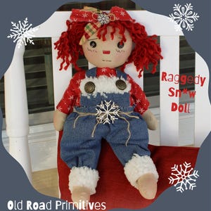 May include: Raggedy Snow Doll handmade with red and white fabric, wearing blue overalls and a white fur trim. The doll is sitting on a red cushion and a white chair. The doll has red curly hair and a red and white plaid bow.